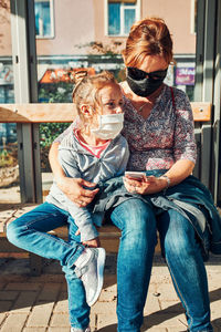 Woman and daughter wearing mask sitting at bus stop