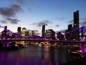 Illuminated bridge over river by buildings against sky at night