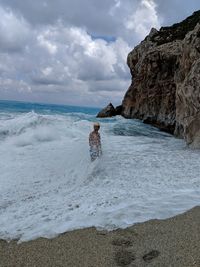 Waves splashing on man standing in sea against cloudy sky