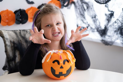 Little girl in costume of witch holding pumpkin jack with candies, celebrating halloween at home