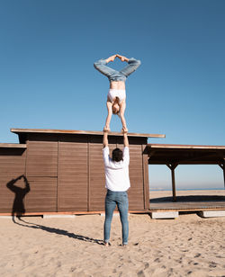 Full body of unrecognizable sportive couple performing hand to hand handstand while standing on sandy shore during training on summer day