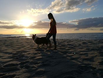 Woman with dog standing at beach against sky during sunset