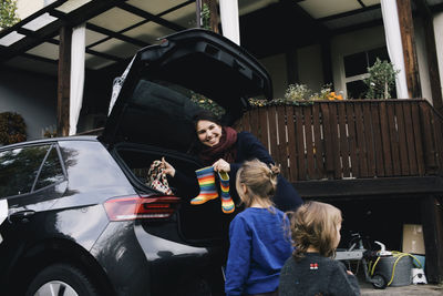 Happy mother with daughters loading boots and bag in trunk of electric car