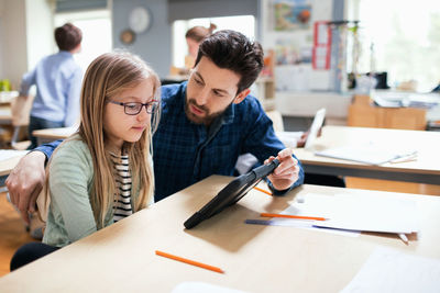 Teacher discussing with student while showing digital tablet at desk in classroom