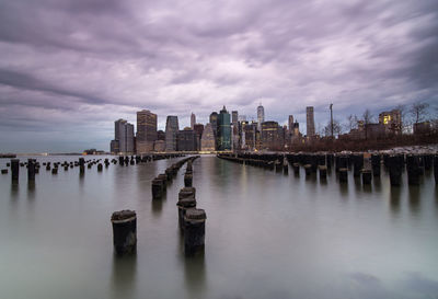 Wooden posts in east river by city against cloudy sky