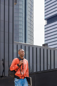 Serious young trendy african american male in stylish outfit with backpack and laptop walking on street near contemporary building on sunny day and looking away