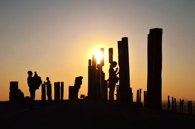 Silhouette people standing by wooden posts against sky during sunset
