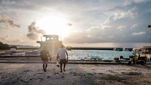 Rear view of men walking at beach against sky during sunset