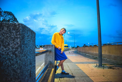 Portrait of smiling young woman standing on footpath against blue sky