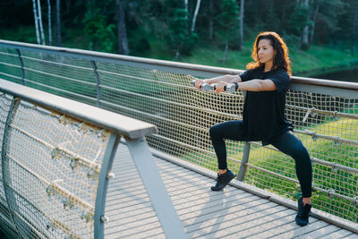 Full length woman lifting dumbbells on footbridge in park