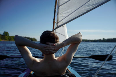 Rear view of shirtless young man sitting on sailing boat in sea against sky