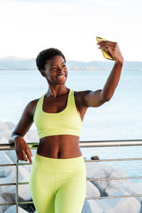 Fit confident african american female in vibrant yellow sportswear focusing on screen and taking selfie with smartphone while standing leaning on metal fence on promenade against blurred seascape