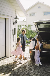 Woman charging electric car while standing with daughters outside house