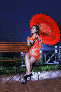 Woman holding umbrella while sitting on bench against sky at night