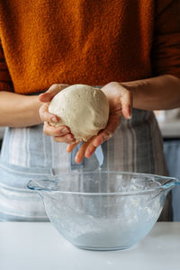 Crop housewife in casual clothes and apron holding dough in hands while standing at table with large glass bowl