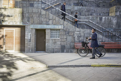 Full length side view of man walking with electric bicycle on footpath in city