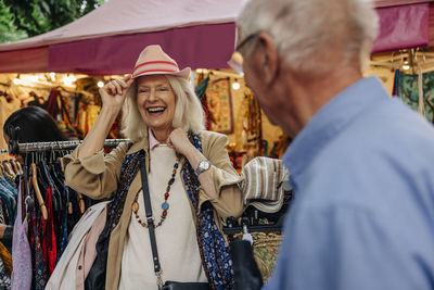 Cheerful senior woman trying on hat by man standing at market