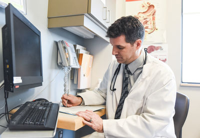 Doctor in a white coat writing in a file at a desk in a clinic room.