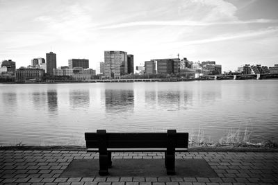 Empty bench by river against buildings in city