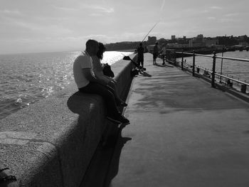 Men sitting on railing by sea against sky