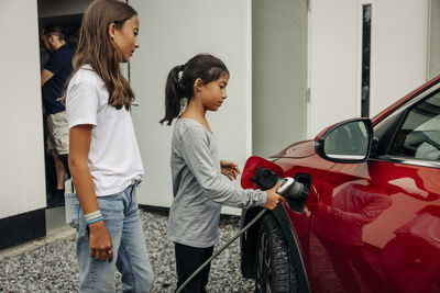 Girl standing next to sister charging electric car near house