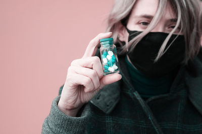 Close-up portrait of woman wearing mask holding capsules against pink background