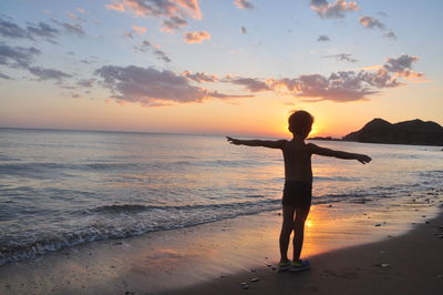 Full length of boy with arms outstretched standing on shore at beach during sunset