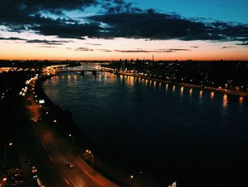 Illuminated city by river against sky at sunset