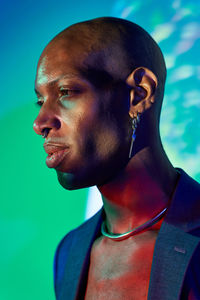 Confident bald african american male model with necklace and earring in trendy outfit standing at wall with glowing lights in modern studio looking away