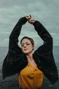 Young woman with arms raised standing at beach against sky