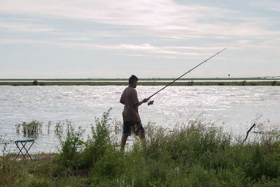 Man with fishing rod walking by sea against sky