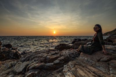 Man sitting on rock by sea against sky during sunset