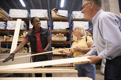 Smiling saleswoman looking at male and female customer holding planks at hardware store