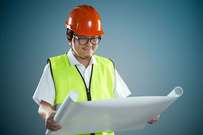 Smiling construction worker reading blueprint while standing against colored background