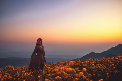 Woman standing by flowering plants against orange sky