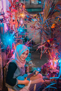 Portrait of smiling young woman wearing hijab playing ukulele by decorations at night