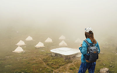 Rear view of young woman standing in foggy weather against tent camping in mountain valley hiking