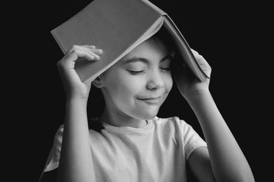 Close-up of smiling girl with eyes closed holding book on head against black background