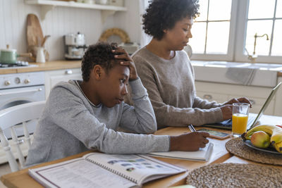 Stressed boy doing homework by mother using laptop at table