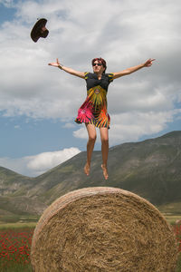 Full length of woman jumping over hay bale against sky