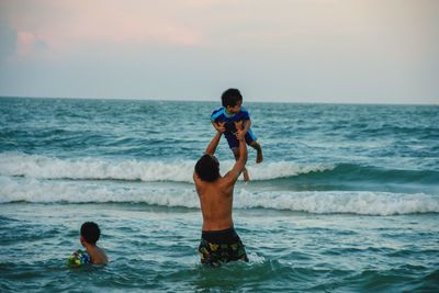 Rear view of shirtless boy lifting brother in sea against sky