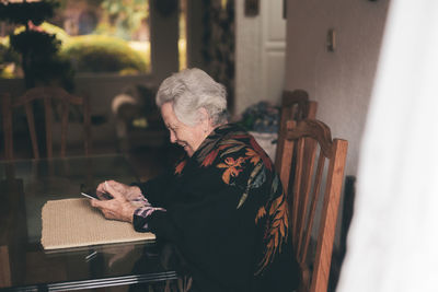 Side view of smiling elderly female wearing warm clothes sitting at table with tablet and cup of tea looking at screen