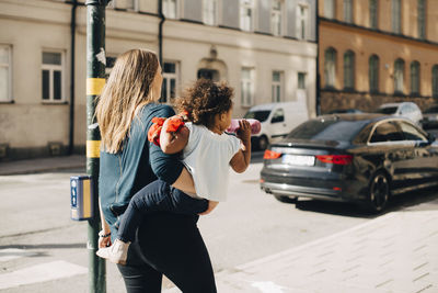 Mother carrying daughter while walking on footpath in city