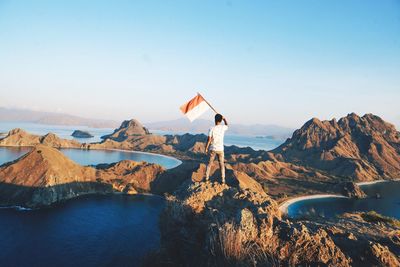 Rear view of man holding flag while standing on rock against clear sky