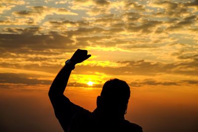 Silhouette man with hand raised against cloudy sky during sunset
