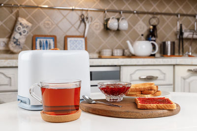 White toaster, cup of black tea, slices of toasted bread, crispy toast with raspberry jam and bowl