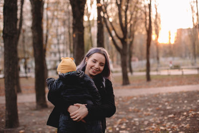 Happy young mother carrying baby son in park in autumn at sunset