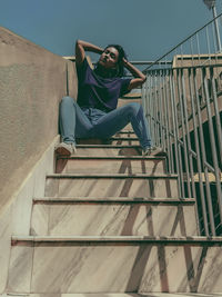 Low angle view of young woman sitting on staircase against sky