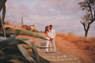 Full length of young couple kissing on steps against cloudy sky