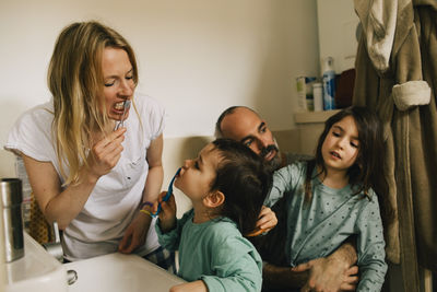 Girl combing brother's hair while he is brushing teeth with mother